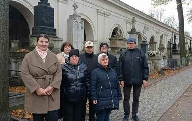 Zdjęie przedstawia grupę os&oacute;b stojących przy nagrobkach w Alei Zasłużonych na Powązkach