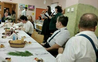 Na zdjęciu grupa kobiet i mężczyzn siedzących przy stole i śpiewających kolędy podczas spotkania wigilijnego 
