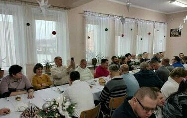 Na zdjęciu grupa kobiet i mężczyzn siedzących przy stole i śpiewających kolędy podczas spotkania wigilijnego 