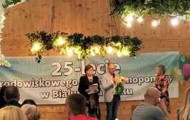 Na zdjęciu dwie kobiety i mężczyzna z mikrofonem w ręku i kwiatami