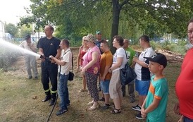 Na zdjęciu grupa os&oacute;b ze strażakiem i mężczyzna lejący mała sikawką strazacką do celu