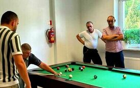 Na zdjęciu czterech mężczyzn przy stole bilardowym, jeden z nich gra w bilarda