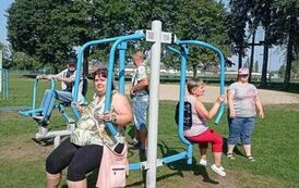 Dwie kobiety i mężczyzna siedzą na urządzeniu fitness zewnętrznym, inna kobieta i mężczyzna stoją z boku