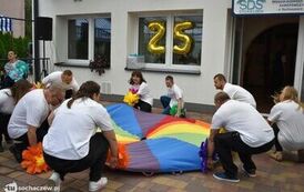 Zdjęcie przedstawia kilka os&oacute;b w białych t-shirtach przykucających podczas zabawy z kolorową chustę animacyjną 