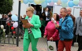 Dwie kobiety i trzech mężczyzna, jedna z nich wręcza upominek mężczyźnie w garniturze; w tle siedzą zaproszeni goście