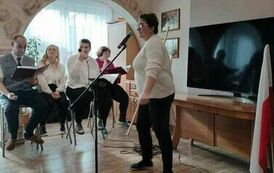 Na zdjęciu kobieta stojąca przy statywie z mikrofonem i śpiewająca pieśni patriotyczne, w tle siedzą  trzy kobiety i mężczyzna