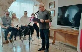 Na zdjęciu mężczyzna stojący przy statywie z mikrofonem i czytający wiersz, w tle na stołkach siedzą odświętnie ubrane trzy kobiety i mężczyzna