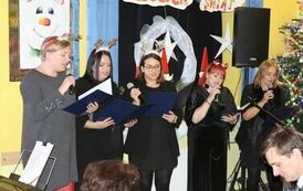 Na zdjęciu, przy udekorowanej choince, stoi pięć kobiet w świątecznych opaskach na głowie śpiewających kolędę