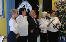Na zdjęciu, przy udekorowanej choince, stoją cztery kobiety i mężczyzna, kt&oacute;rzy śpiewają kolędę