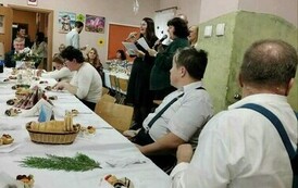 Na zdjęciu grupa kobiet i mężczyzn siedzących przy stole i śpiewających kolędy podczas spotkania wigilijnego 