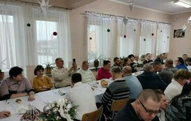 Na zdjęciu grupa kobiet i mężczyzn siedzących przy stole i śpiewających kolędy podczas spotkania wigilijnego 