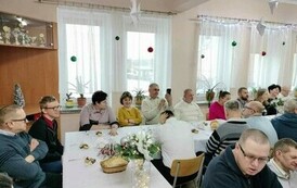 Na zdjęciu grupa kobiet i mężczyzn siedzących przy stole i śpiewających kolędy podczas spotkania wigilijnego 
