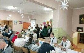 Na zdjęciu grupa kobiet i mężczyzn siedzących przy stole i śpiewających kolędy podczas spotkania wigilijnego 