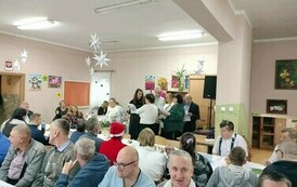 Na zdjęciu grupa kobiet i mężczyzn siedzących przy stole i śpiewających kolędy podczas spotkania wigilijnego 