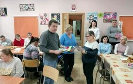 Zdjęcie przedstawia kobietę czytającą życzenia i mężczyznę przekazującego laurkę drugiej kobiecie. Obok stoją i siedzą kobiety i mężczyźni.