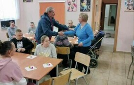Zdjęcie przedstawia mężczyznę składającego życzenia kobiecie. Obok siedzą kobiety i mężczyźni.