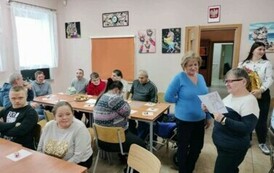 Na zdjęciu stoją dwie kobiety. jedna z nich trzyma w ręku kartkę z życzeniami. Obok przy stołach siedzą kobiety i mężczyźni.